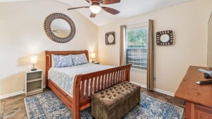 A bedroom with a wooden bed, a ceiling fan, a large mirror, a window with blinds, and a bedside table with a lamp. - 17007 Firenzo Ave 3 Bedroom Home (Panama City Beach)