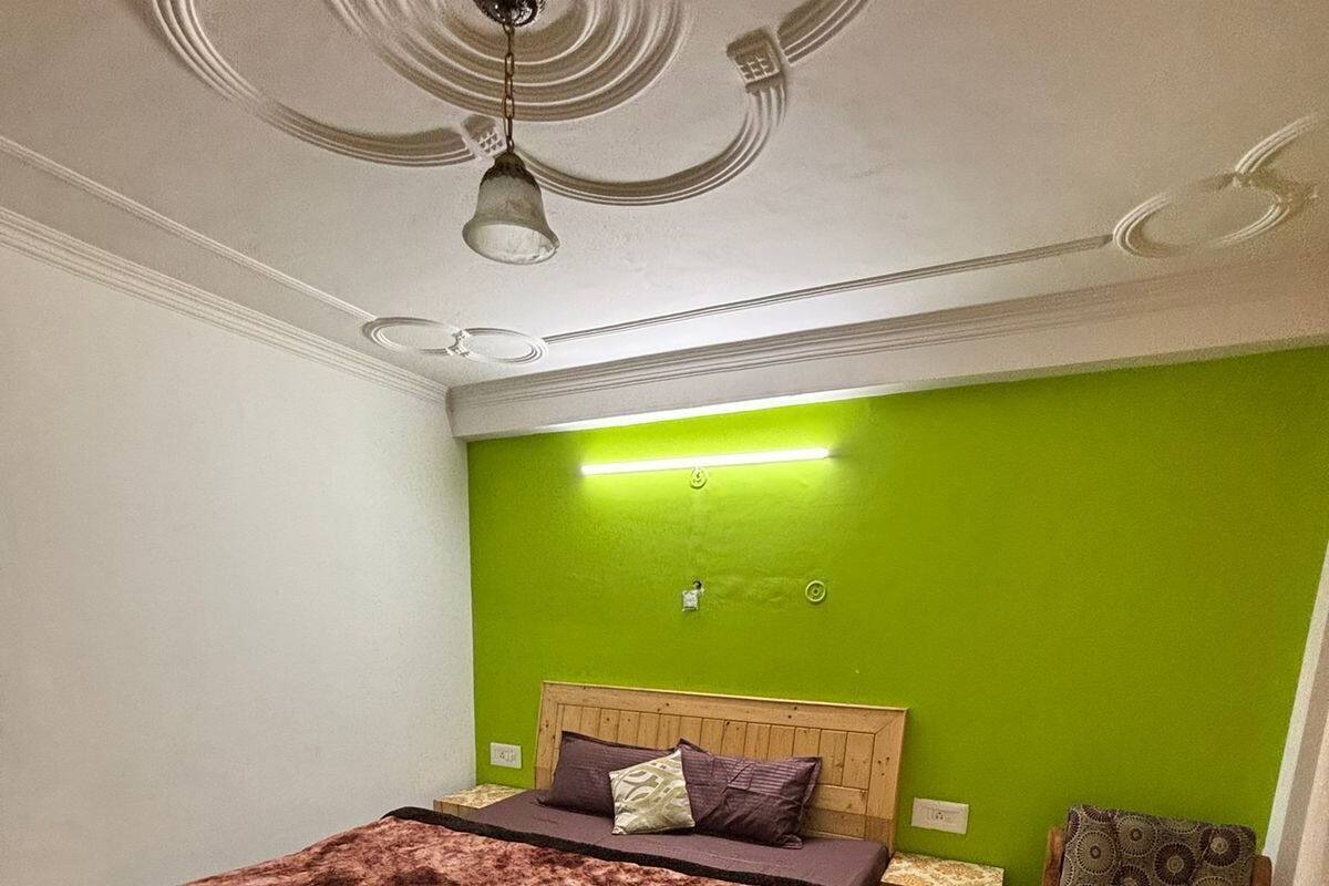 Un dormitorio con cama de madera, paredes verdes, alfombra estampada, mesita de noche de madera y una silla.