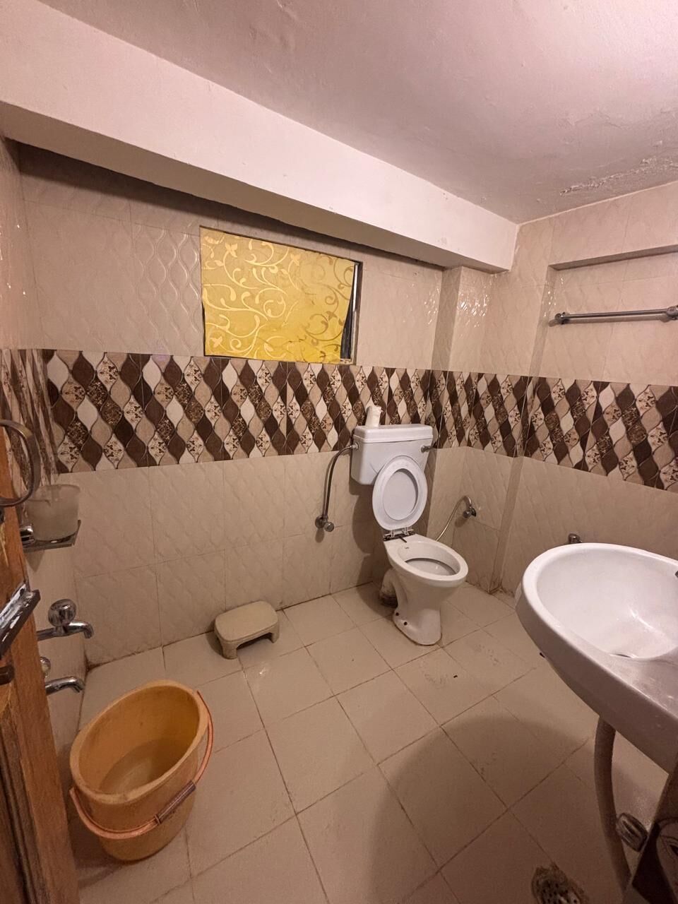 A bathroom with a toilet, bidet, and sink. There is a bucket on the floor and a towel rack on the wall.