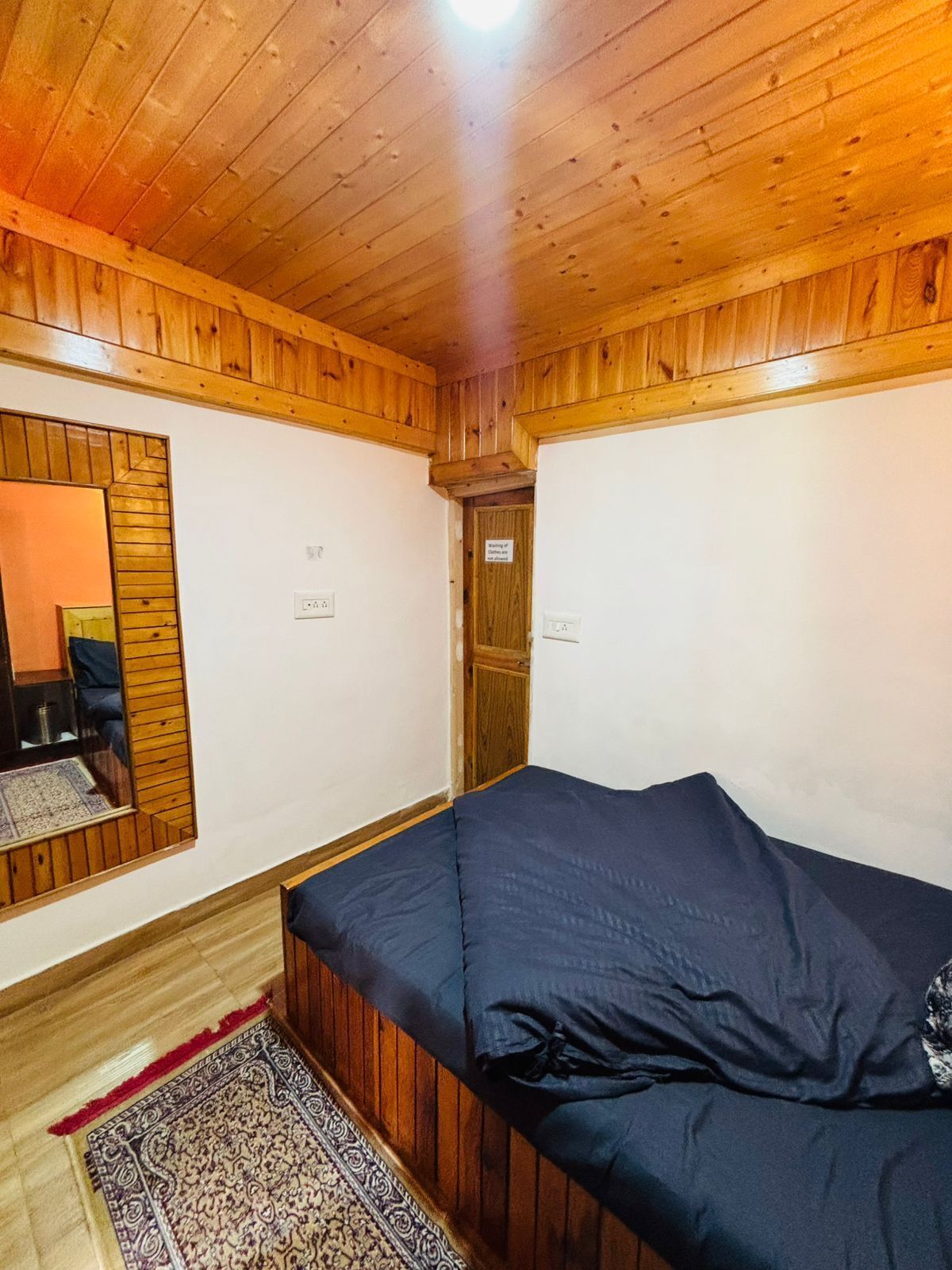 Quarto com paredes e teto de madeira, cama com roupa de cama escura, um espelho e uma porta com uma placa.