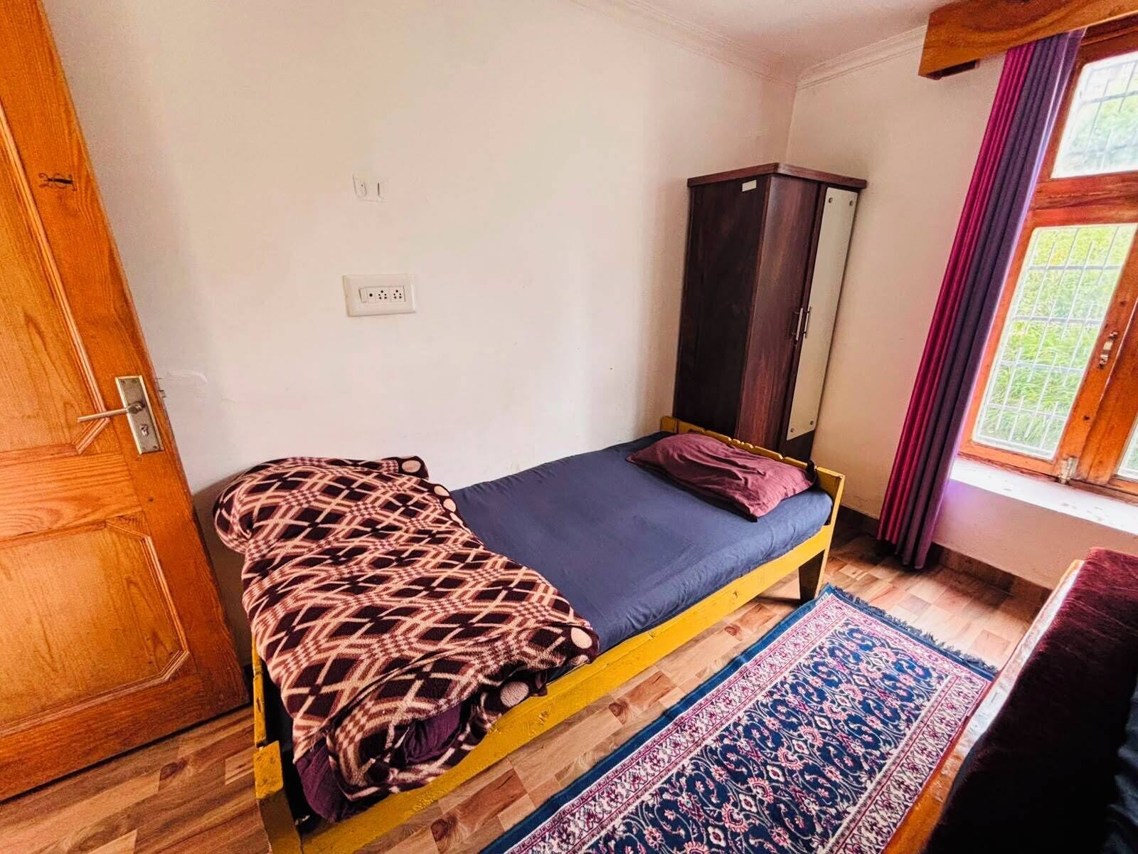 Ein kleines Zimmer mit einem Einzelbett, einem Holzschrank, einer gemusterten Tagesdecke und einem blauen Teppich.
