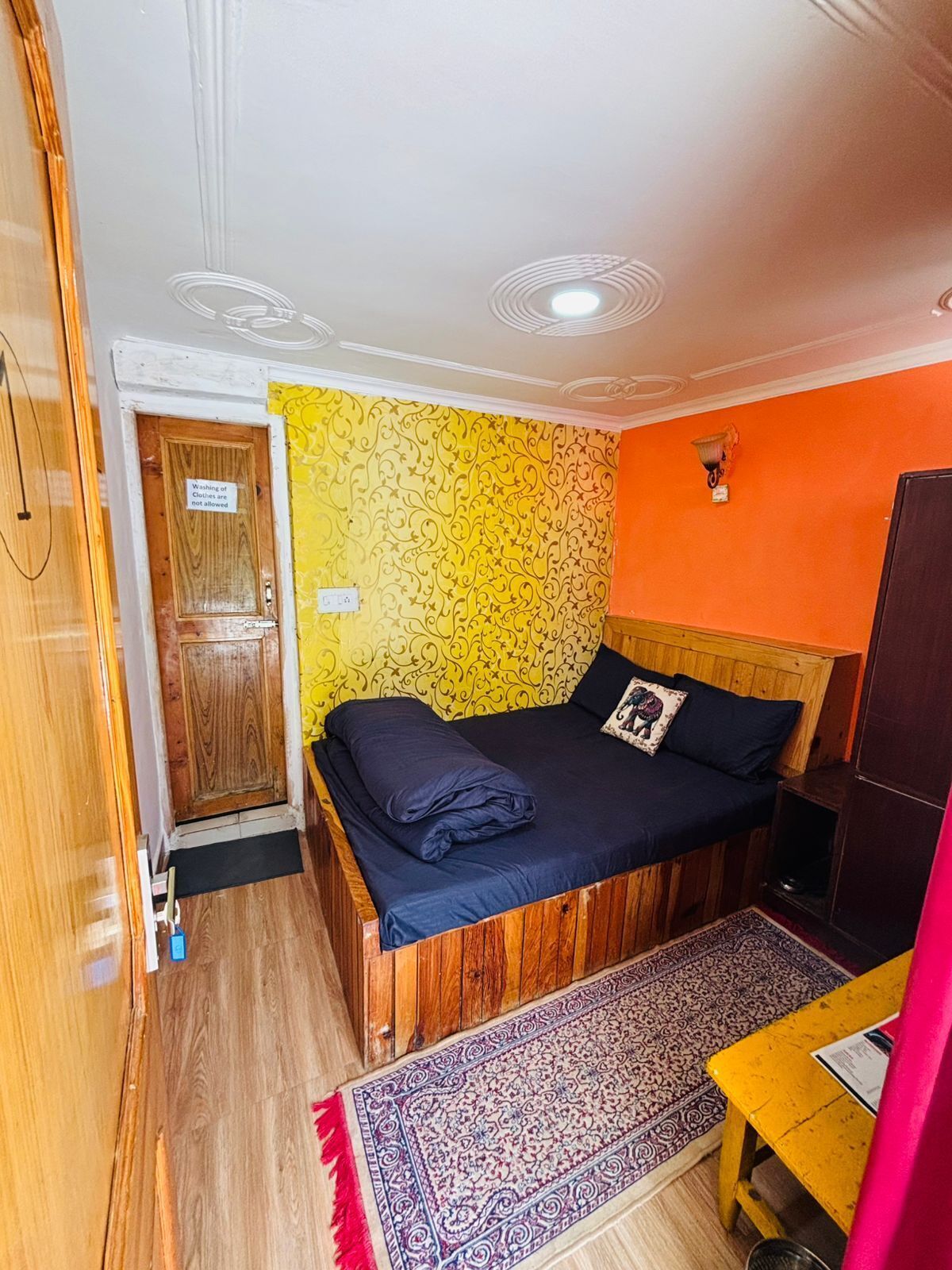 A small, compact room with a bed, a wooden wardrobe, a door with a sign, and a small table with a chair.