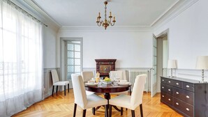 A dining room with a round table, four chairs, a chandelier, a cabinet, and a window with curtains.