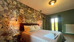 Une chambre avec un papier peint à motifs floraux, une tête de lit en bois, un lit avec une literie blanche, une table de chevet avec une lampe, et une vue sur l’extérieur grâce à une porte coulissante.