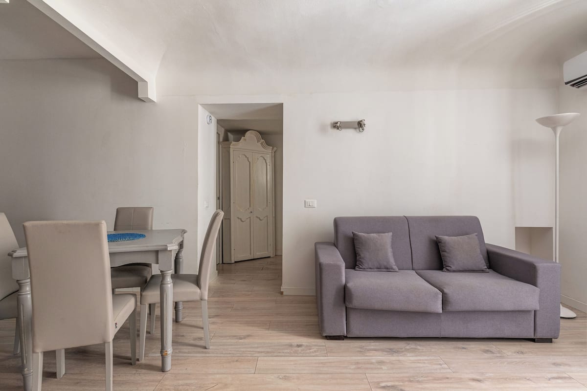 A modern living room with a grey sofa, a dining table with chairs, and a hallway leading to a door.