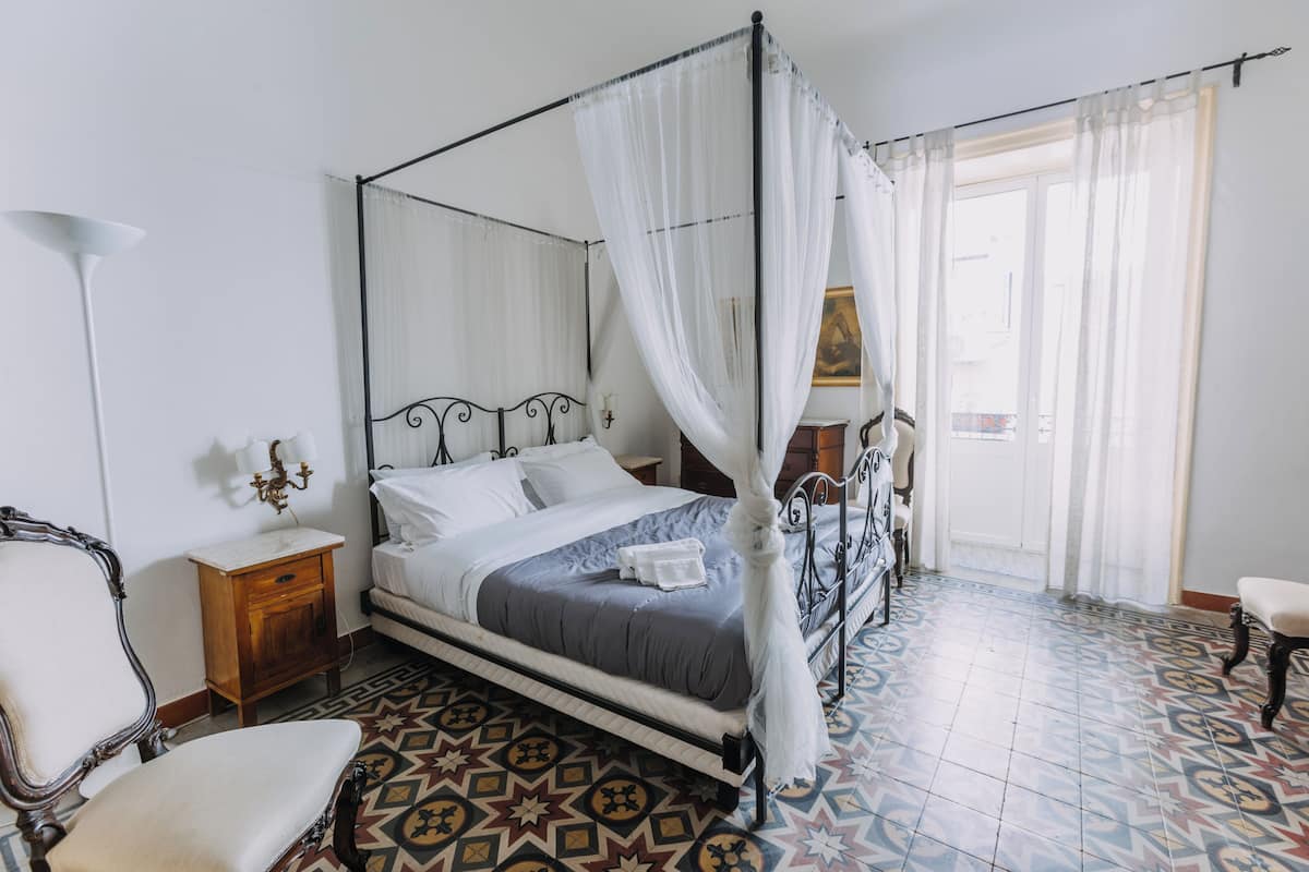 A four-poster bed with a canopy, a bedside table with a lamp, a chair, and a patterned floor.