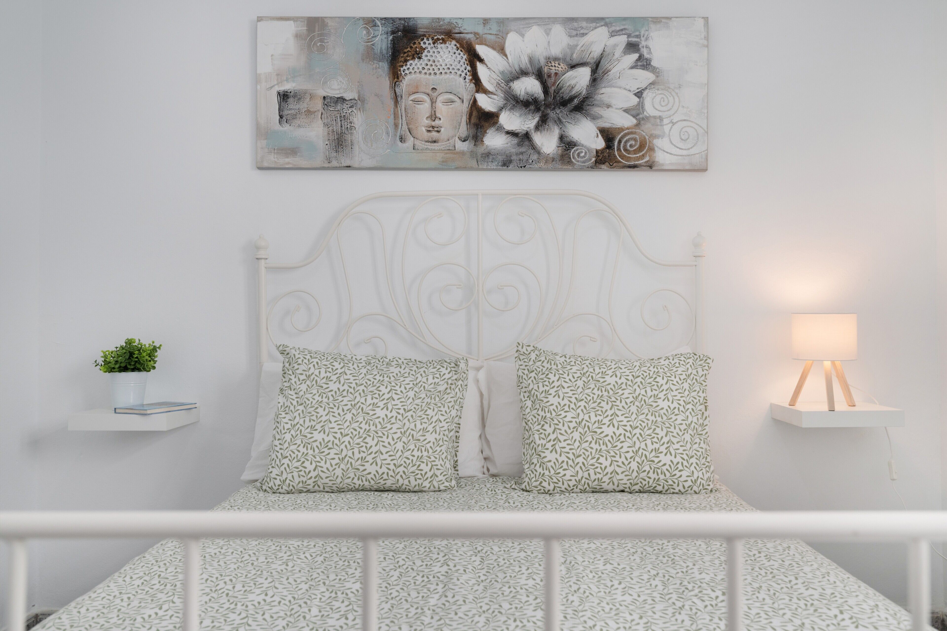 Ein Bett mit weißem Metallgestell, zwei Kissen mit Muster, eine kleine Zimmerpflanze auf dem Nachttisch und ein an der Wand hängendes Kunstwerk.
