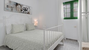 Ein Schlafzimmer mit einem Bett, einem Nachttisch, einem Wandbild, einem Fenster mit grünen Rollläden und einem weißen Vorhang.