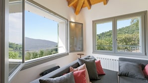 Un salon moderne avec un canapé gris, des coussins rouges et gris, et de grandes fenêtres donnant sur un paysage verdoyant et des montagnes.