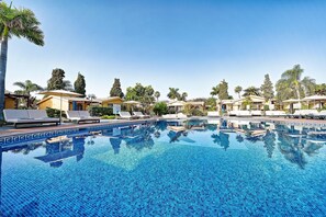 Une piscine entourée de palmiers et d’immeubles, avec des chaises longues et des parasols.