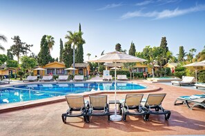Un espace piscine aménagé avec des chaises longues, des parasols et une piscine à l’eau cristalline, le tout entouré de palmiers et de bâtiments.