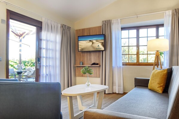 Habitación de hotel con sofá, una mesita con un jarrón, televisión en la pared y ventana con cortinas.