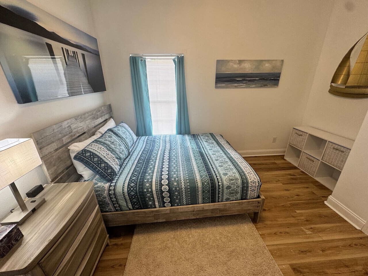 A bedroom with a bed, a nightstand, a lamp, a window with curtains, and a wall-mounted shelf.
