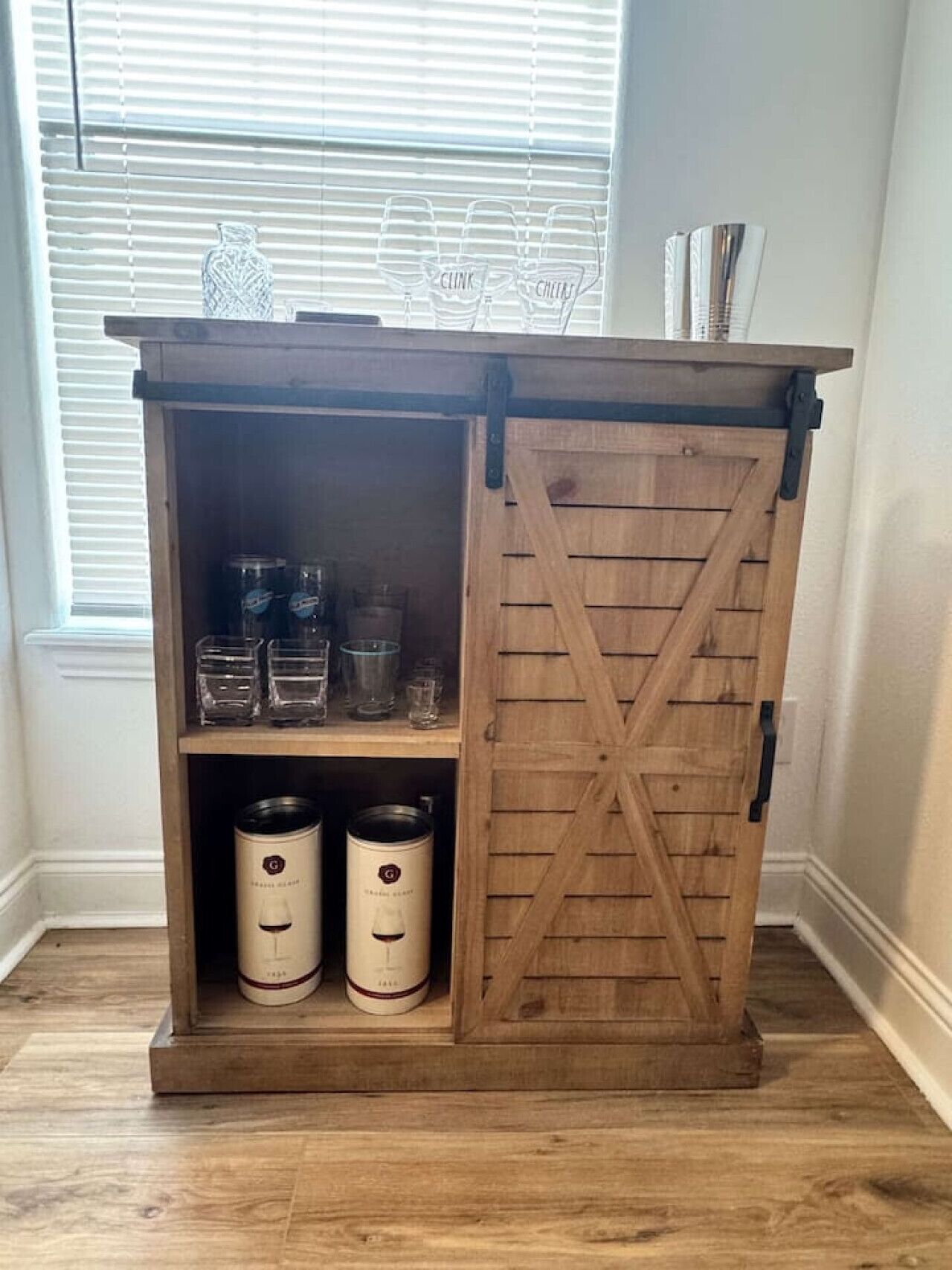 A wooden cabinet with a barn-style door, shelves with glassware, and two white containers.