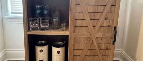 A wooden cabinet with a barn-style door, shelves with glassware, and two white containers.