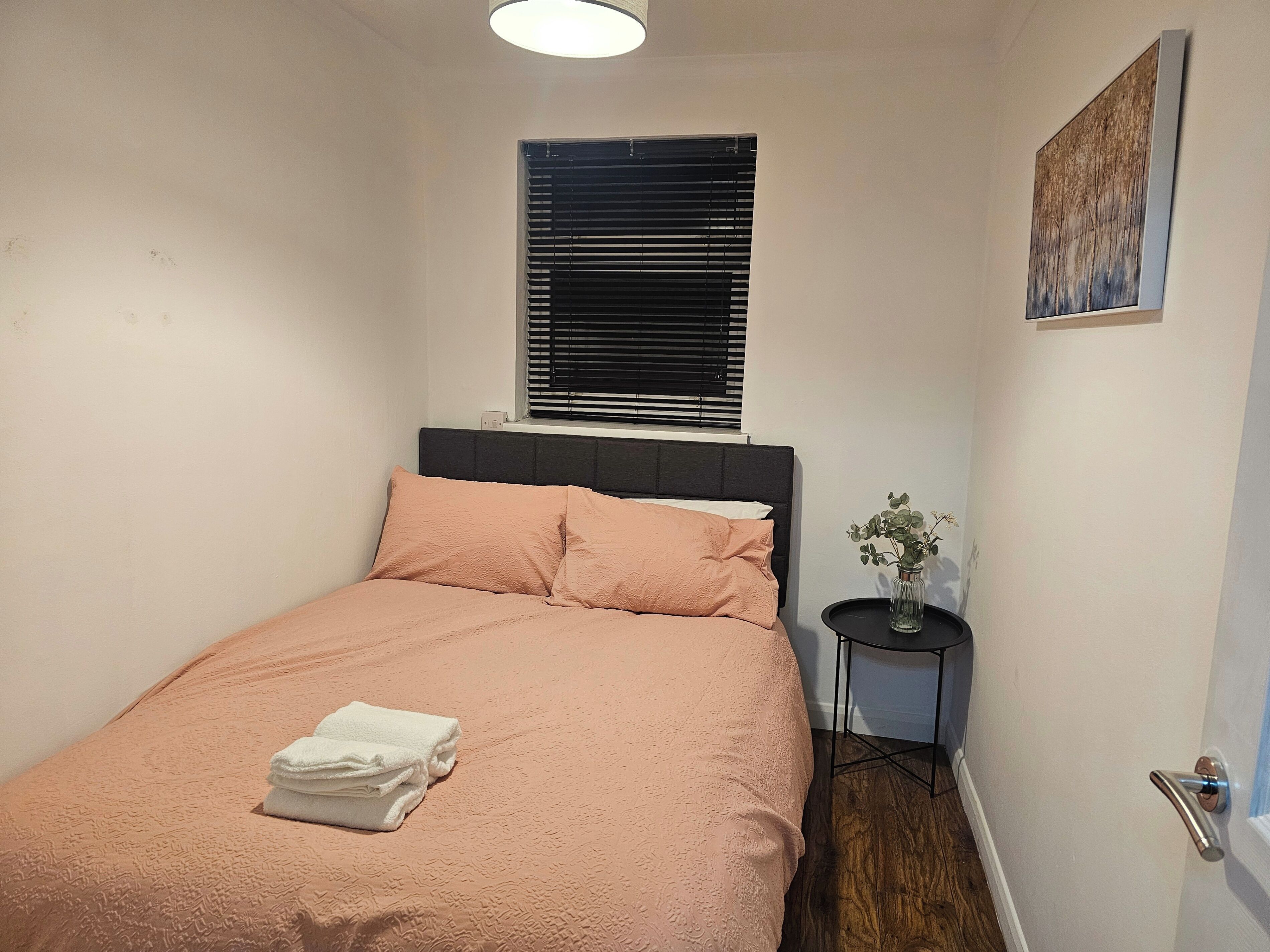 A neatly made bed with a pink bedspread and white pillows, a small bedside table with a vase of flowers, and a window with blinds.