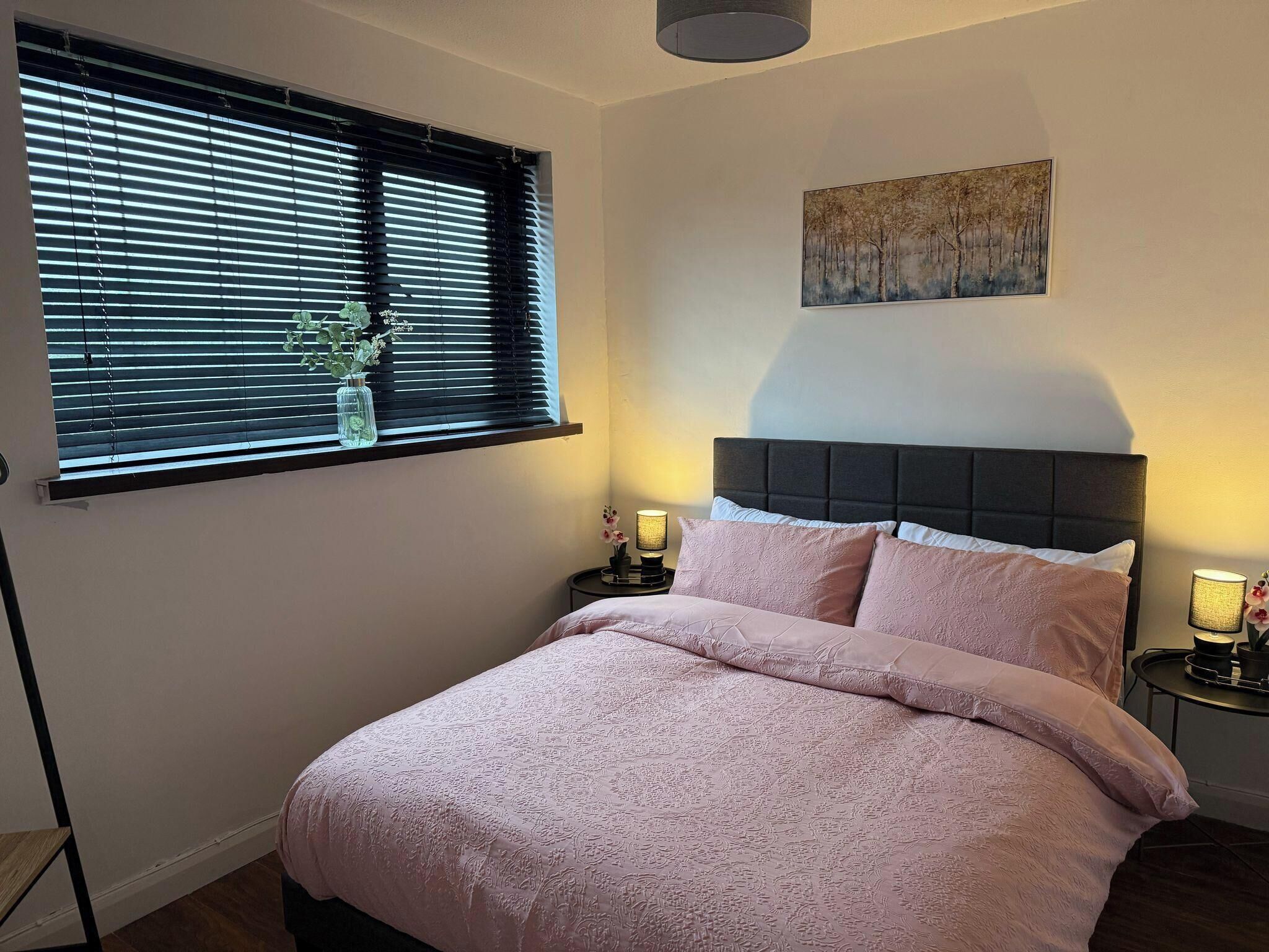 Un dormitorio con cama, ventana con persianas, mesita de noche, lámpara y un cuadro en la pared.