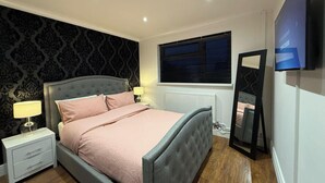 Ein Schlafzimmer mit Bett, Nachttischen, einer Lampe, einem Fenster mit Jalousien, einer Heizung und einem an der Wand montierten Fernseher.