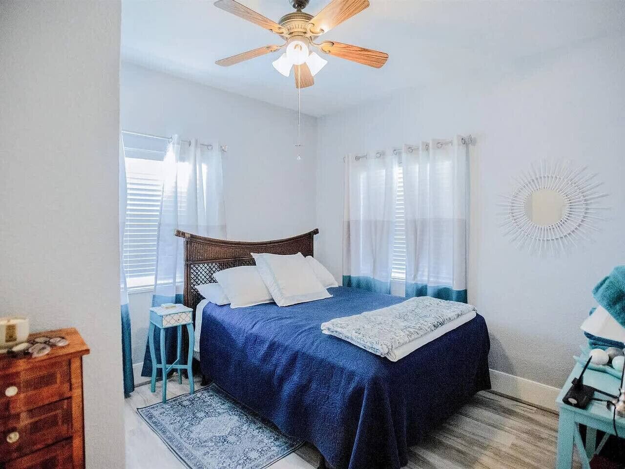 A bedroom with a bed, a wooden nightstand, a blue chair, a ceiling fan, and a wall-mounted mirror.