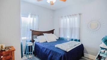 A bedroom with a bed, a wooden nightstand, a blue chair, a ceiling fan, and a wall-mounted mirror.