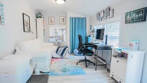 Une pièce moderne et bien éclairée, avec un canapé blanc, un bureau avec un ordinateur, une petite kitchenette et un rideau bleu.