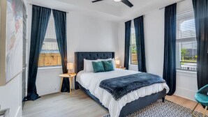 Un dormitorio con una cama, dos mesitas de noche, un ventilador de techo y ventanas grandes con cortinas.
