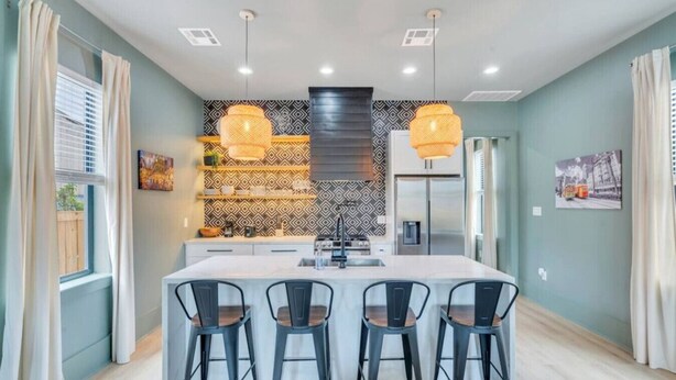 Una cocina moderna con una isla central, taburetes negros tipo barra, iluminación colgante y un salpicadero con diseño.
