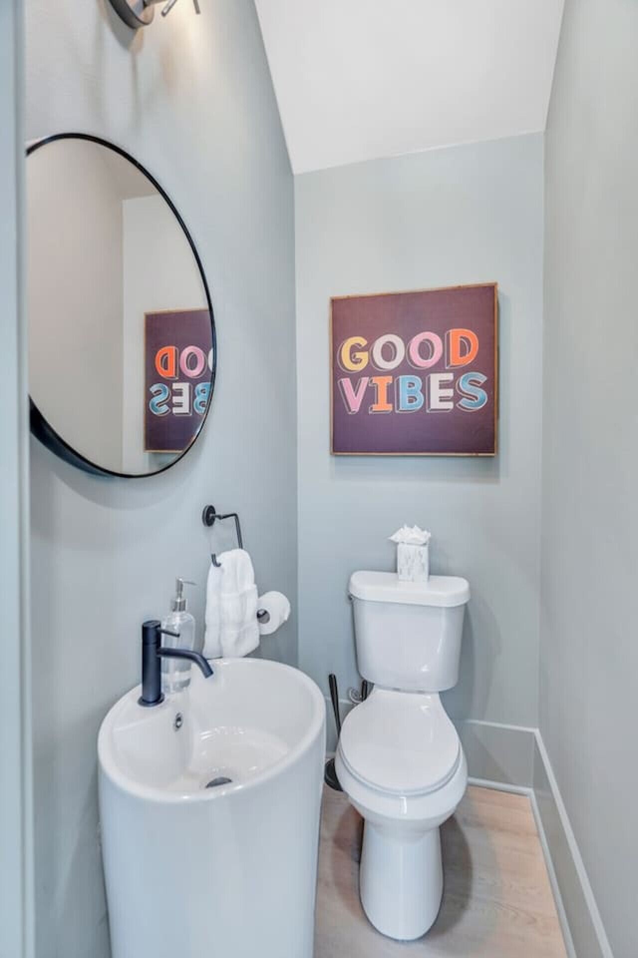 Une salle de bain avec un lavabo, des toilettes et un miroir. Un panneau est fixé au mur, sur lequel on peut lire : « BONNE AMBIANCE ».