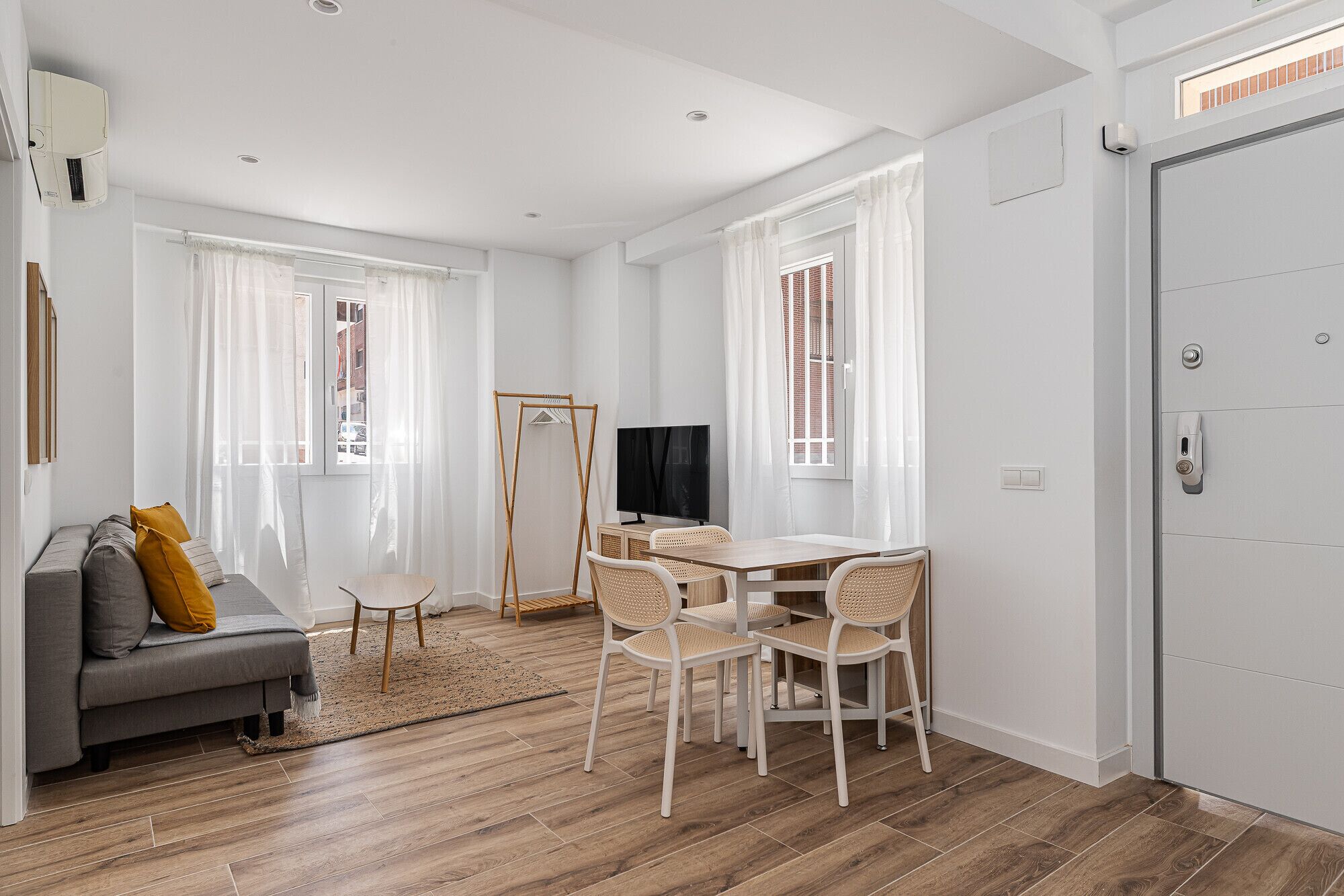 Una sala moderna con un sofá gris, una mesita redonda pequeña y un televisor. Hay dos ventanas con cortinas blancas, un piso de madera y una puerta blanca.