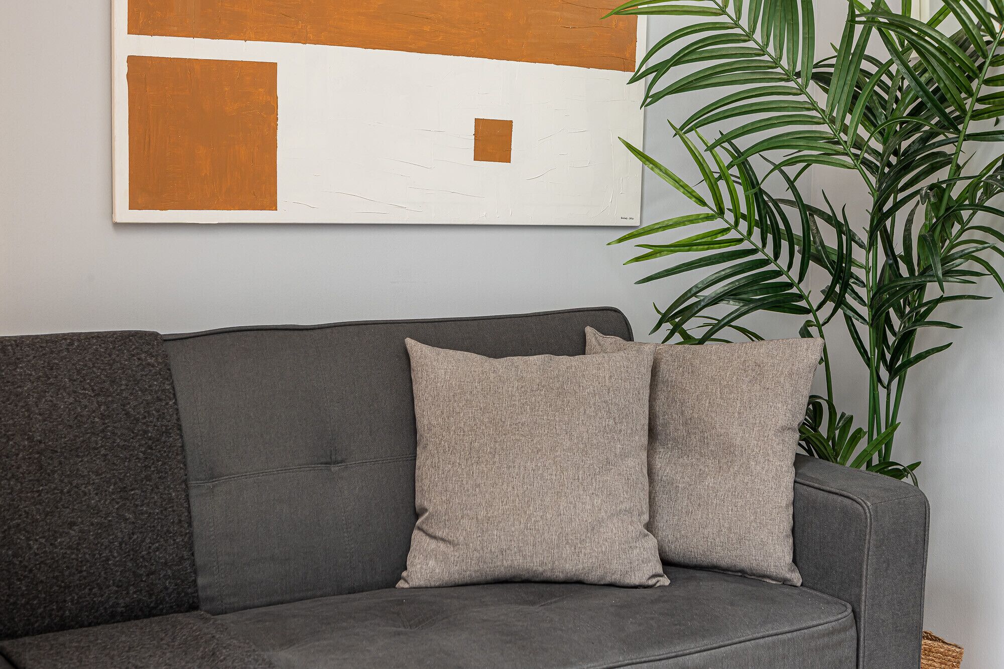 Ein graues Sofa mit zwei beigen Kissen, ein großes abstraktes Gemälde an der Wand und eine grüne Pflanze neben dem Sofa.