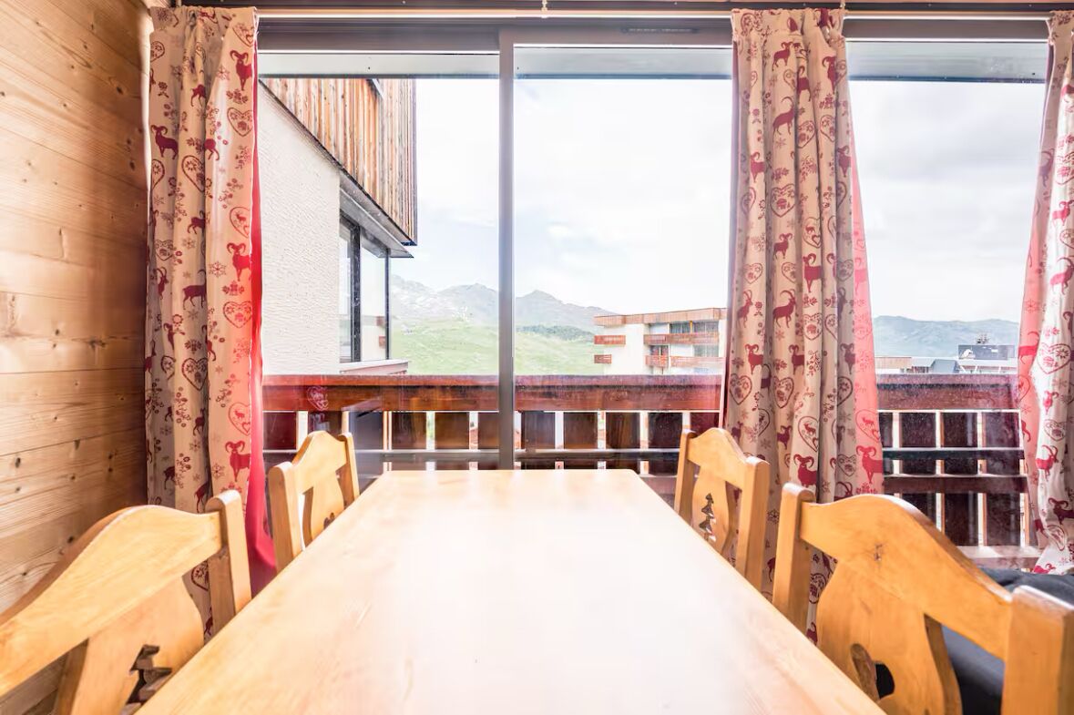 Ein Essbereich mit Holztisch und -stühlen, großen Fenstern mit gemusterten Vorhängen und Blick auf Gebäude und Berge.