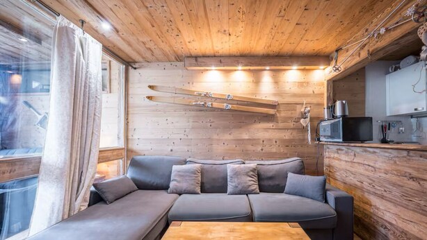 Een gezellig interieur van een chalet met een houten plafond, een grijze bank, een houten bijzettafeltje en ski's die aan de muur hangen.