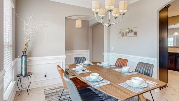 Comedor con una mesa de madera dispuesta para seis personas, sillas blancas, un candelabro y un florero con ramas sobre una mesita auxiliar.