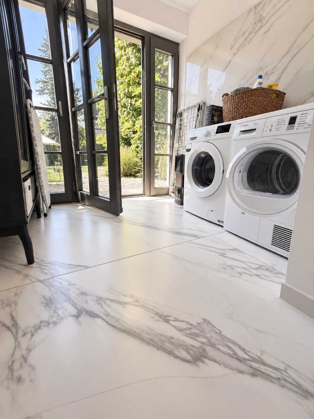 A laundry room with two front-loading washing machines, a large window with a view of greenery, and a marble floor.