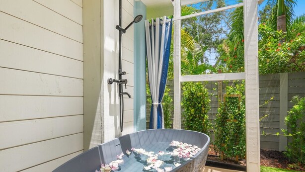 A freestanding bathtub with rose petals floating on the water, a showerhead, and a white pergola with a view of a garden.