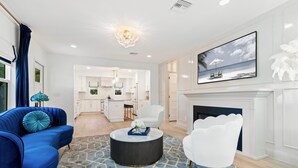 A modern living room with a blue sofa, white armchair, and a round coffee table. A fireplace and a large framed picture are visible.
