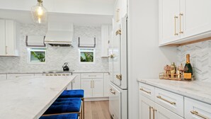 A modern kitchen with white cabinetry, gold handles, and a marble countertop. There are two blue cushioned stools at the island.