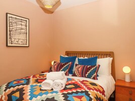 A neatly made bed with a colorful quilt, two pillows, and rolled towels. A framed artwork is on the wall, and a bedside lamp is on the nightstand.