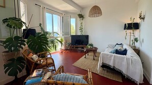Una sala de estar con televisor, muebles de mimbre, una planta colgante y una cama con edredón blanco.