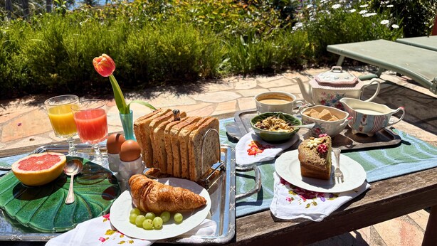 Desayuno con tostadas, croissant, uvas y jugo en una mesa al aire libre con vista al mar.