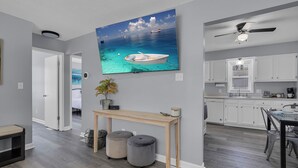 A modern kitchen with white cabinets, a dining area with a table and chairs, and a living space with a TV displaying a boat on water. - Walk to Clark Lake + Beach Bar, Firepit, Porch, Sleeps 8 (Clarklake)