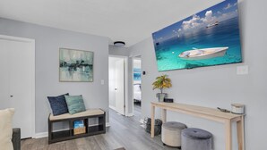 A hallway with a bench, a painting, a console table, and a television displaying a boat on water. - Walk to Clark Lake + Beach Bar, Firepit, Porch, Sleeps 8 (Clarklake)