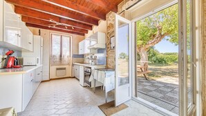Une cuisine avec des meubles blancs, une bouilloire rouge et une vue sur un arbre à travers une porte ouverte.