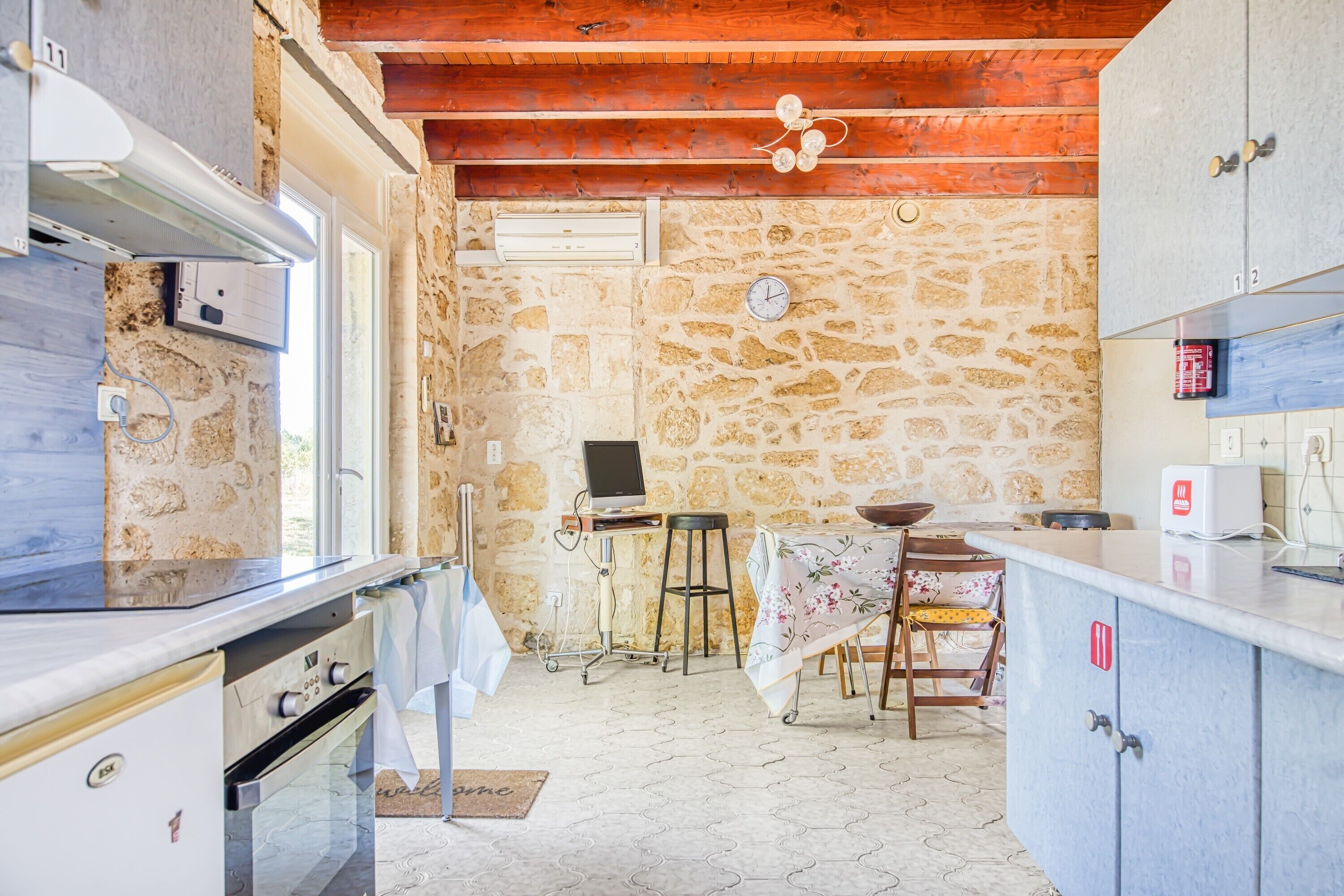 Une cuisine avec des murs en pierre, des poutres en bois au plafond, une table à manger avec des chaises et un petit téléviseur.