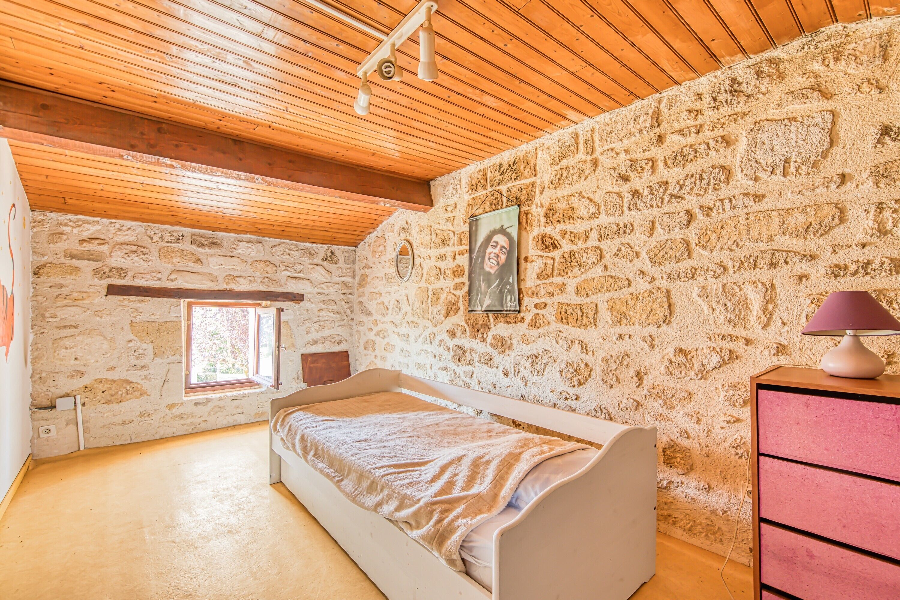 Een slaapkamer met een stenen muur, houten balken in het plafond, een bed, een dressoir en een raam.