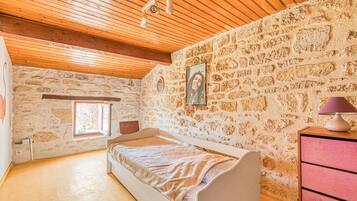 Een slaapkamer met een stenen muur, houten balken in het plafond, een bed, een dressoir en een raam.