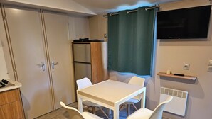 A compact room with a white table and four chairs, a small kitchenette with a refrigerator, and a TV mounted on the wall. - Bungalow Privas (AZALÉE (Unknown)