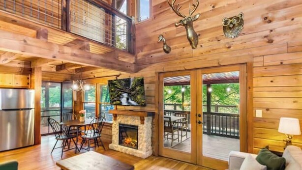 A spacious wooden cabin interior with a stone fireplace, dining area, and a balcony with a view of the outdoors.