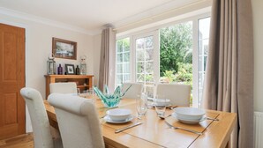 A dining area with a wooden table set for four, chairs, a glass bowl centerpiece, and a view of greenery outside. - Lorien (Cheltenham)