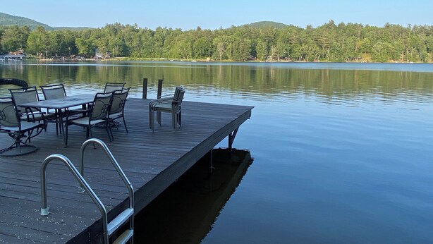 A wooden dock with chairs and tables, extending over calm water with a forested hill in the background.